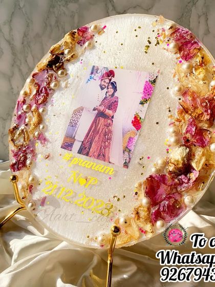 A beautiful round resin plaque with preserved varmala flowers, pearls, and a central photo. It sits elegantly on a golden stand, making it a perfect decorative piece for a side table or shelf.