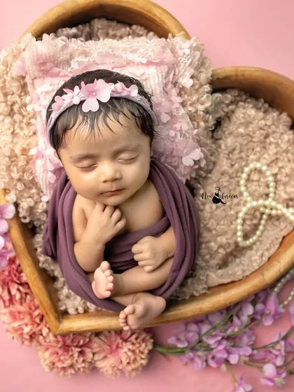 This precious newborn girl is nestled in a heart bowl, wrapped in a lovely purple swaddle. The floral details and soft textures create a beautiful, feminine portrait.