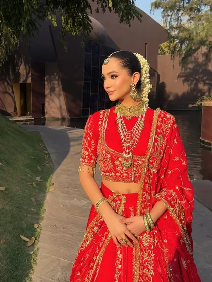 A side profile capturing the elegance of a traditional bride. The styling focuses on the harmony between the red lehenga, the heavy jewelry, and the classic floral hair adornment.