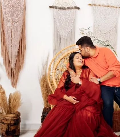 A candid, happy moment captured during a studio maternity shoot. The couple shares a laugh in a comfortable, boho-inspired setting with natural textures and plants.