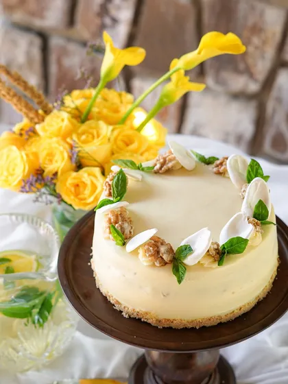A beautiful cheesecake decorated with walnuts and fresh basil, set against a backdrop of yellow flowers. This is another one of our celebration cakes available for special occasions like Mother's Day.