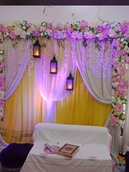 An intimate indoor Haldi decoration for a home setting. This arrangement uses layers of yellow and sheer white drapes, a floral border of pink and white flowers, and hanging lanterns to create a warm and cozy ambiance.