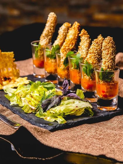 An elegant presentation of sesame-crusted fish fingers served in shot glasses with a savory dip, accompanied by a fresh green salad.