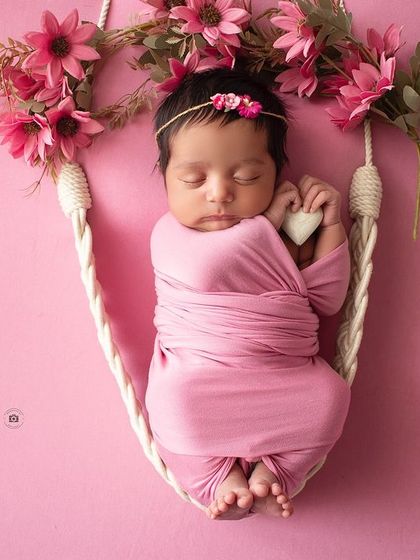 A newborn baby girl, wrapped snugly in pink, sleeps on a floral swing prop.