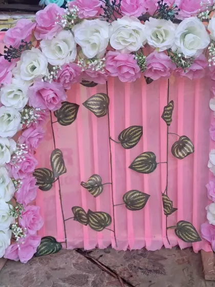 A simple and elegant backdrop with an arch of pink and white artificial roses. The pink fabric background is decorated with artificial betel leaves, offering a clean and natural-looking design.