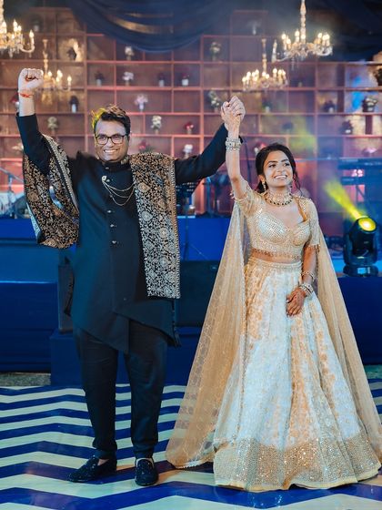 The couple takes a bow on stage after their sangeet performance, their hands raised in celebration.
