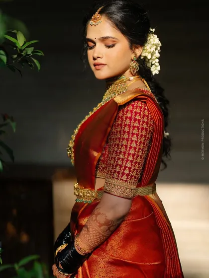 Bride Archana radiates elegance in a rich orange Kanjeevaram saree. The ensemble is completed with an intricate hand-embroidered blouse that adds a touch of modern allure to her traditional look.