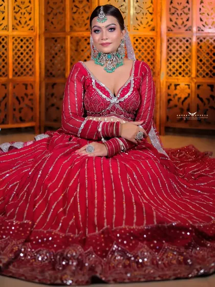 A full seated shot showing the spread of the red lehenga against a traditional carved wooden screen.