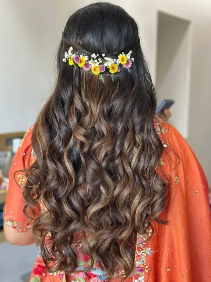 A beautiful half-updo with voluminous curls and highlights, accented with a fresh flower clip for a mehendi look.