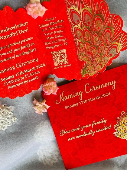 A vibrant and traditional naming ceremony invitation in auspicious red. The card features a beautiful gold foil peacock motif and a unique scalloped, tri-fold design that makes it feel extra special. It also includes a QR code for easy navigation to the venue.