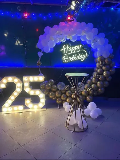 A club-style 25th birthday setup. This decoration uses a partial balloon arch in gold and white against a dark backdrop, with a neon sign and marquee numbers.