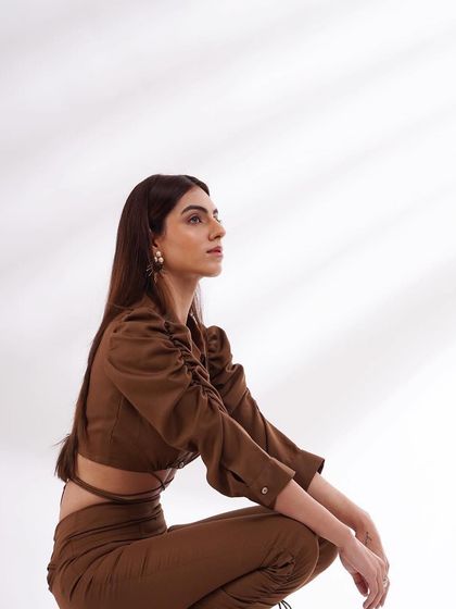 A profile shot of a model in a brown ruched crop top and trousers. The clean, minimalist background keeps the focus entirely on the garment's texture and fit.
