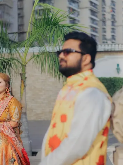 A candid shot of the couple during their Haldi event. The groom in the foreground with the bride smiling in the background creates a lovely, layered composition.