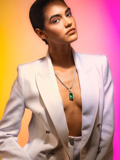 A bold and powerful portrait using colored gels. The contrast between the sharp white suit, the vibrant background, and the striking emerald necklace creates a high-fashion, editorial feel.