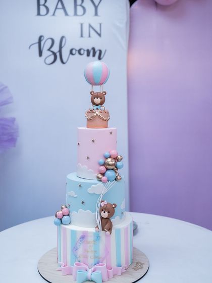 A stunning three-tiered cake for a "Baby in Bloom" themed baby shower. The pastel colors, hot air balloon, and teddy bear decorations are captured to remember the beautiful details of the celebration.
