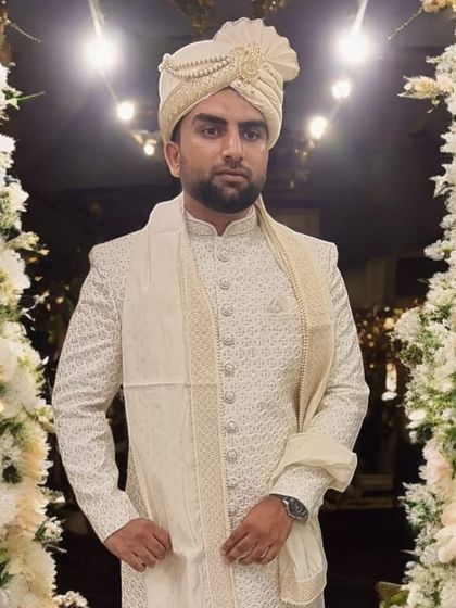 A happy client looking dapper on his wedding day. He wears a custom-fitted, intricately patterned off-white sherwani paired with a matching turban and stole, embodying pure elegance.