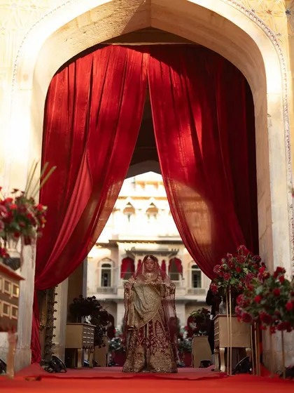 A dramatic bridal entry through heavy red velvet curtains, creating a sense of royal theatre and anticipation. The architecture of the palace is used to frame this unforgettable moment.