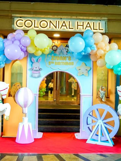 A whimsical pastel carnival entrance with carousel horses, hot air balloon props, and a beautiful balloon arch over the doorway.