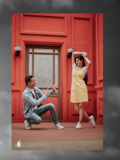 A playful and stylish pre-wedding shot against a vibrant red wall, capturing the groom-to-be's fun proposal-like pose.