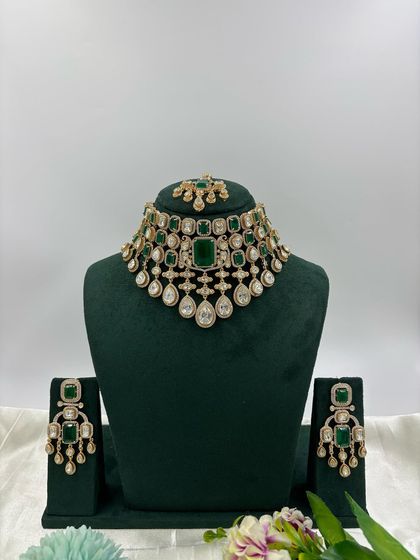 A regal, high-neck choker set featuring a large emerald-green stone at its center. The design is complemented by hanging pearls and matching earrings, perfect for creating a statement bridal look.