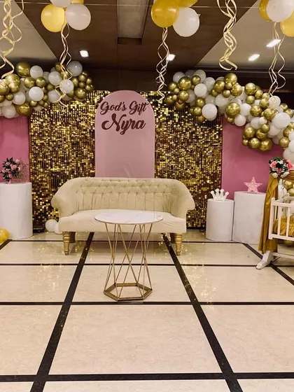 A glamorous baptism decor setup in pink, white, and gold. The stage features a gold shimmer backdrop, balloon arches, and a decorated cradle for the baby.