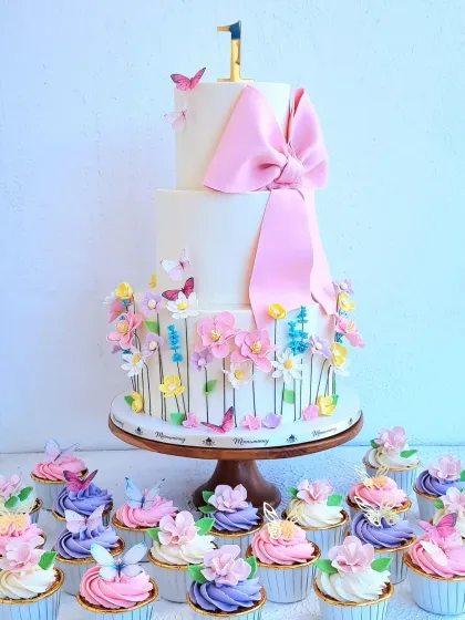 A beautiful two-tier first birthday cake with a garden theme, complete with a large pink fondant bow. This set includes matching floral cupcakes, showing how I can create a cohesive and elegant dessert table for your celebration.