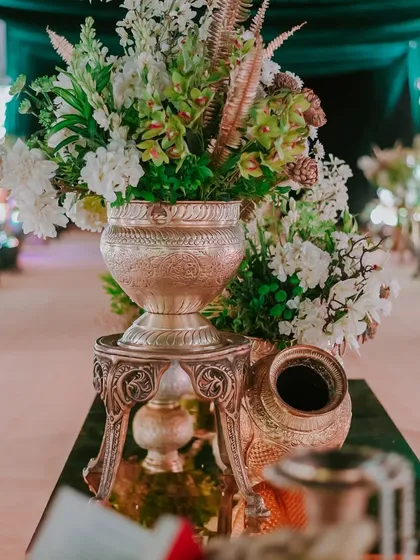 A floral arrangement in a silver vase, combining white blooms with interesting green textures and gilded ferns, sits on a mirrored table, reflecting the surrounding opulence.
