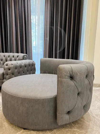This living room features a curved, tufted Chesterfield sofa in grey, paired with a matching circular ottoman. The set creates a cohesive and elegant look, grounded by dark marble nesting tables.