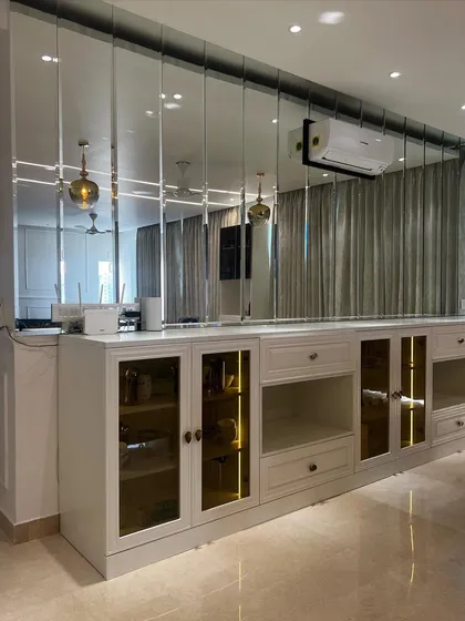 This custom bar and crockery unit in Gurgaon features a mirrored back, glass-front cabinets with internal lighting, and a mix of open and closed storage, all framed by a large mirror wall to enhance the room's spaciousness.