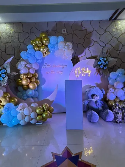 A dreamy moon-and-stars themed baby shower decoration. This "Oh Baby" setup features a crescent moon backdrop, a large plush teddy bear, and a celestial-inspired balloon arrangement in blue, white, and gold.