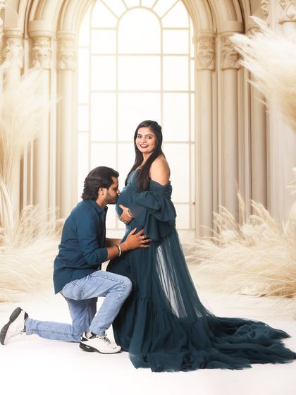 A classic kneeling kiss to the baby bump, captured in a grand studio setting with architectural elements and pampas grass. The flowing gown adds a touch of ethereal beauty.