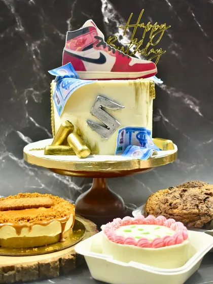 I can create a complete dessert spread for your event. This table features a custom sneaker-themed drip cake as the centerpiece, surrounded by a creamy Biscoff cheesecake, giant chocolate chip cookies, and a personal-sized bento cake, ensuring there's a delicious treat for every guest.