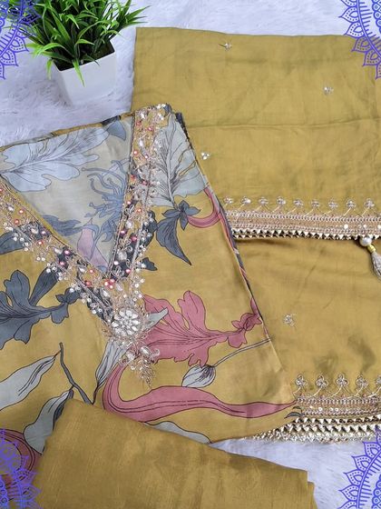 A fancy dress material in a mustard yellow shade with a digital floral print. The neckline is embellished with mirror work, and the dupatta has a delicate embroidered border.