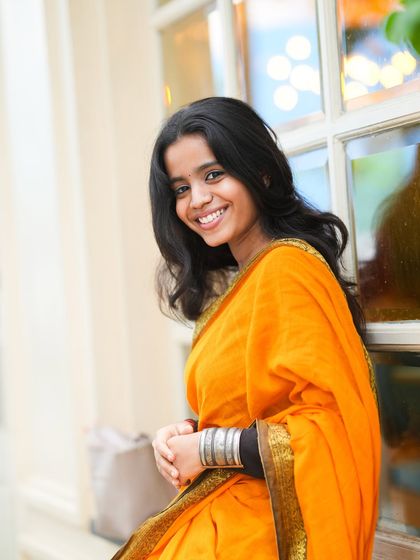 A bright and cheerful portrait of a young woman in a vibrant orange saree, captured in a natural, outdoor urban setting, perfect for a fashion or lifestyle portfolio.