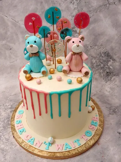 This "He or She, Can't Wait to See" cake features adorable blue and pink teddy bears, colorful lollipops, and a fun drip effect.