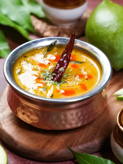 A bowl of tangy and sweet Kairi Dal, a raw mango lentil curry, garnished with a red chili. The rustic wooden board and fresh mango leaves create a scene that feels like a summer day in an Indian home.