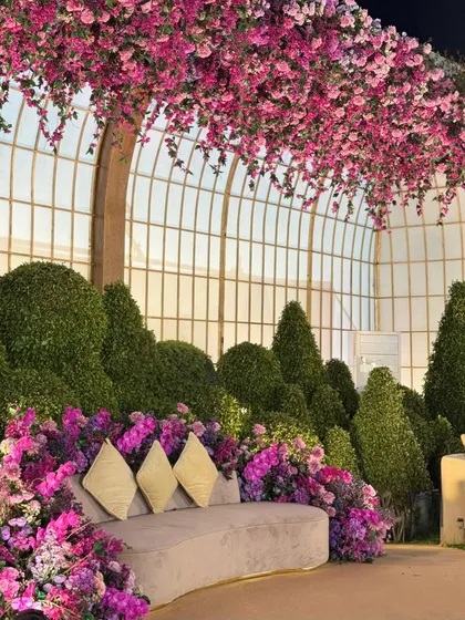 A luxurious curved sofa set amidst a landscape of sculpted topiaries and vibrant flowers. The large arched windows in the background add to the feeling of being in a grand conservatory.