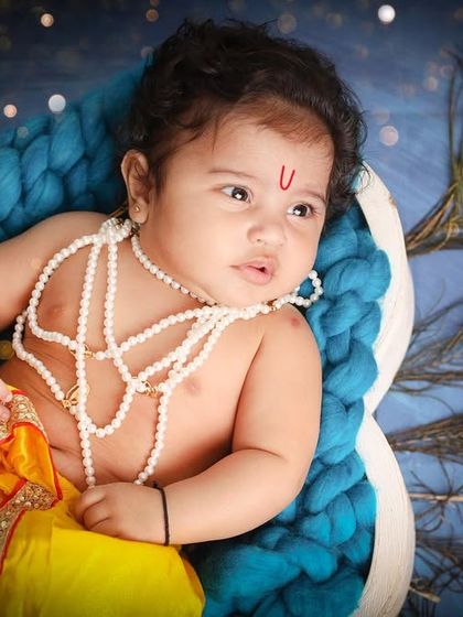 A serene portrait of a baby boy dressed as Krishna, nestled in a basket with peacock feathers. This is a beautiful way to celebrate a baby's first Janmashtami.