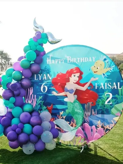 An outdoor Little Mermaid birthday setup. This decoration features a circular backdrop with Ariel, a balloon garland, and a mermaid tail accent.