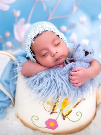 A sweet newborn holding a tiny blue teddy bear, posed in a watering can prop for a whimsical garden-themed shoot.