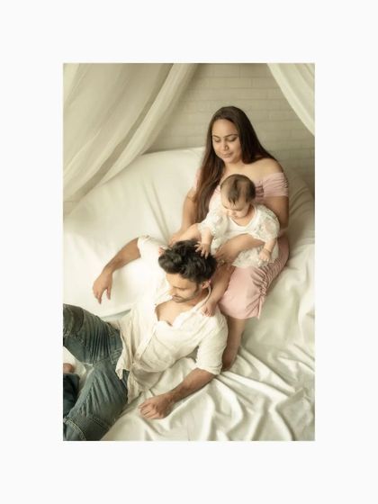 A full family portrait showing the unbreakable bond between parents and their child. The baby playfully sits on her father's head as he lies down, a moment of pure, unscripted family fun.