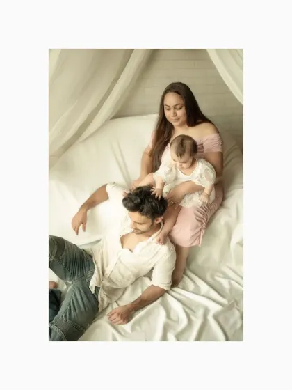 A playful moment captured on a soft, white bed setup in the studio. The child gently plays with her father's hair as her mother looks on, creating a candid and loving family portrait that feels natural and relaxed.