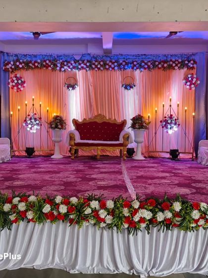 A full view of a wedding event stage with a floral border along the front. The combination of warm and cool uplighting on the backdrop creates a dynamic look, highlighting the central red sofa for the couple.
