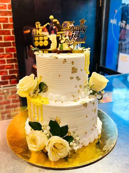 A "Finally Got Engaged" two-tier cake with a cheerful yellow drip, fresh yellow roses, and delicate gold leaf accents.