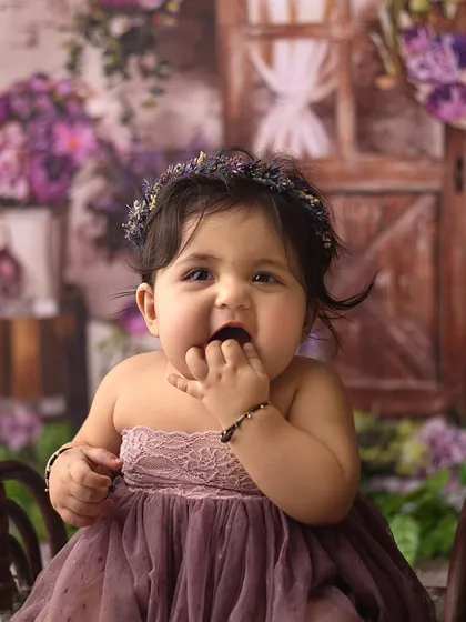 Cuteness has just been overloaded. This little beauty looks so adorable with her fingers in her mouth, surrounded by purple flowers.