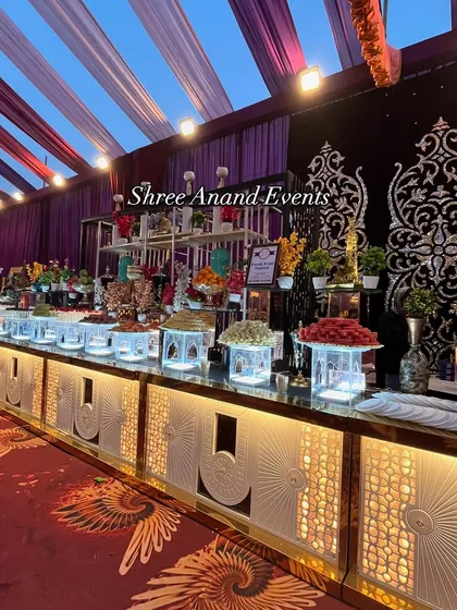 A magnificent dessert and fruit counter that serves as a visual masterpiece. The display is built on illuminated platforms with intricate silver and gold backdrops, featuring everything from fresh fruit to traditional Indian sweets.
