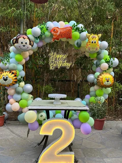 A fun and colorful jungle animal theme for a second birthday. This outdoor setup uses a circular balloon arch decorated with friendly animal foil balloons like a monkey, tiger, and giraffe, complete with a neon sign and a large marquee number '2'.