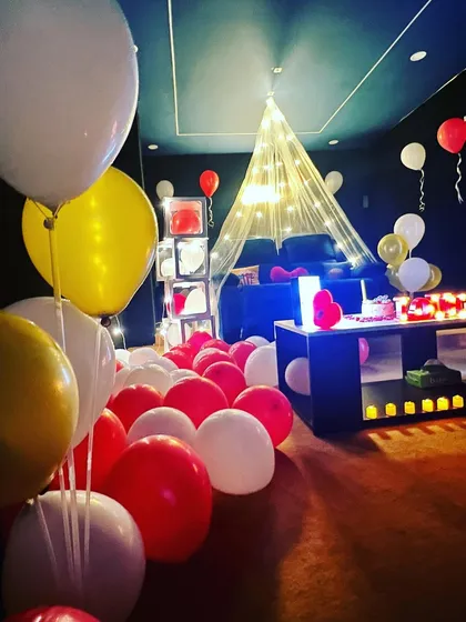 A full view of a romantic setup. The room is filled with red, white, and yellow balloons, with a cozy canopy over the recliner, creating an immersive and celebratory environment.
