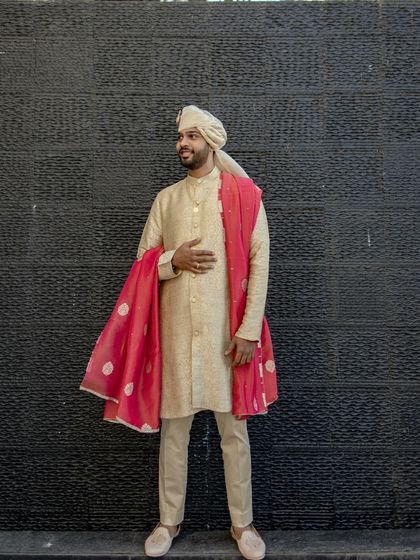 A stylish portrait of the groom in a beige sherwani with a contrasting red dupatta. His confident stance against a simple black wall makes the outfit pop.