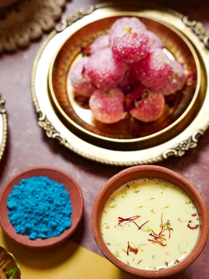 A close-up of traditional Holi sweets, including pink Chum Chums and a bowl of Thandai. The earthy terracotta bowls and bright blue gulal create a festive and rustic composition.